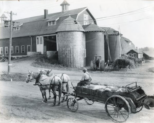 Elevated view of a man driving a manure spreader pulled by a team of horses. In the background is a large barn with three silos, a tractor, wagons and an automobile. A sign on a power line post reads: "Auto Tour".