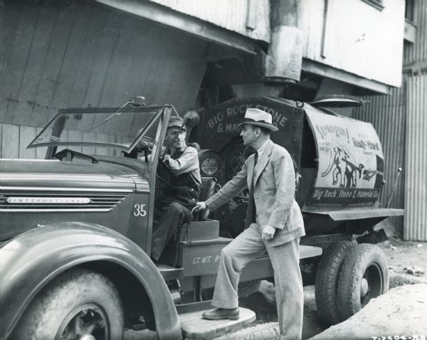 A man identified as R.S. Wilson, Jr., who is wearing a hat and a suit, gives directions to a truck driver working for Big Rock Stone & Material Co. The original caption reads: "R.S. Wilson, Jr. gives directions to driver of Big Rock Stone and Materials Company. Mixed concrete delivery unit, one of 25 trucks in the fleet, one of the largest fleets of its kind in the South." The truck is a concrete mixer, and it is parked near an industrial building underneath a chute.