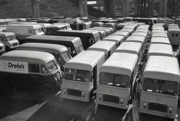 Metro trucks and Metro-Mites parked bumper to bumper in an outdoor lot at the Metropolitan Body company, an International Harvester subsidiary. The truck at left has "Drake's Cake" and "Drake Bakeries Incorporated" painted on its side.