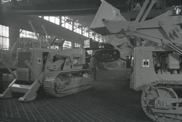 Workers steady a crawler tractor (TracTracTor) as it is moved by a ceiling-mounted conveyor at an International Harvester factory, possibly Tractor Works.