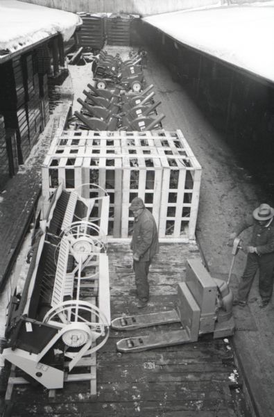 Elevated view of a factory worker using a forklift on a loading dock to move a field harvester on a pallet at International Harvester's McCormick Works. Another man stands near the forklift, and there is snow on the roof of the loading dock.
