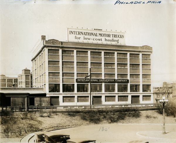 Exterior view of International Harvester's Philadelphia branch house. Automobiles are parked in a cul-de-sac in front of the building; a sign reading: "International Motor Trucks for Low-Cost Hauling" stands atop the roof.