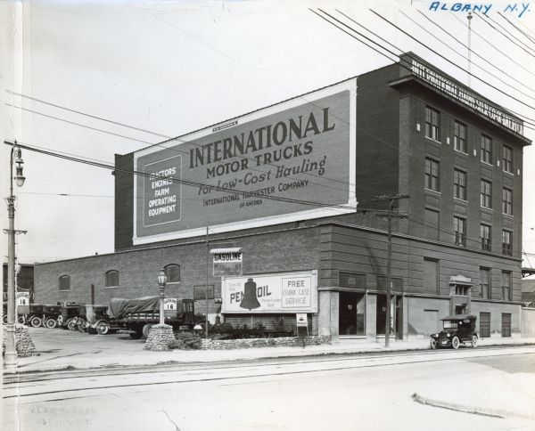 Automobile and trucks are parked alongside the International Harvester branch building in Albany. A sign painted on the upper side of the building reads, "International Motor Trucks for Low-Cost Hauling. International Harvester Company of America. Tractors. Engines. Farm Operating Equipment." Advertisements for Pennzoil and MotoZest are on the lower side of the branch building.