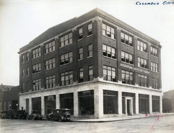 Exterior view of International Harvester Company's Columbus branch house. Several automobiles are parked alongside the curb. A railroad car is behind the building on the right.