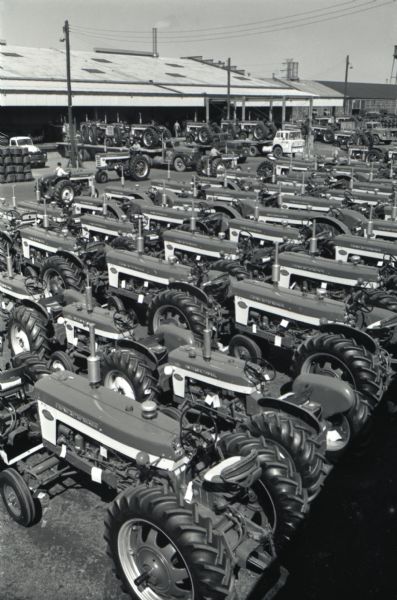 Elevated view of rows of Farmall tractors lined up in a lot outside International Harvester's Farmall Works. Original caption (with magazine article) reads: "Impressive but hardly novel is the sight of Farmalls rolling out of Rock Island, Illinois, by the trainload and truckload. From the day last summer when the new line was introduced, the tractor builders at Farmall Works have had to step lively to keep up with customer demand. Since then their ranks have been swelled by several hundred and their daily production rate has risen 34$. The order bank remains substantial. At Louisville Works, home of smaller Farmalls and utility tractors, schedules have been boosted by 25%. Despite a 65-day strike, IH farm tractor sales are running ahead of the pace of '58."