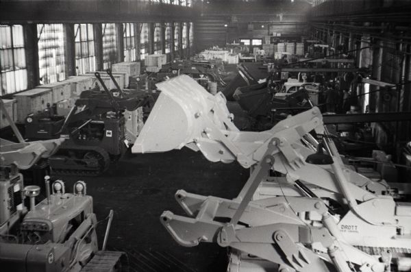 Elevated view of multiple rows of crawler tractors lined up inside a factory - most likely Tractor Works. A group of men are working in the far background. A tractor in the foreground has a Drott loader.