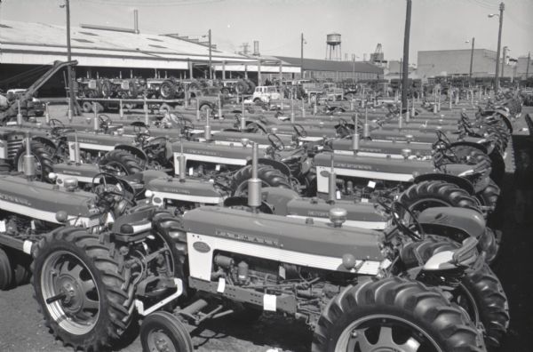 Rows of Farmall tractors stand outside International Harvester's Farmall Works. The factory site includes Industrial buildings and a water tower.