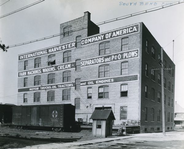 Exterior rear view of International Harvester's South Bend branch building. A railroad car stands on tracks that run past the building at the loading dock. The painted sign reads: "International Harvester Company of America. Farm Machines, Wagons, Cream-Separators and P&O Plows. Motor Trucks, Oil Tractors and Oil Engines."