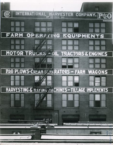 Exterior view of International Harvester's St. Louis branch building. The painted signs read, "International Harvester Company of America Inc. Farm Operating Equipments. Motor Trucks — Oil Tractors & Engines. P&O Plows — Cream Separators — Farm Wagons. Harvesting & Haying Machines — Tillage Implements." The tops of railroad cars are in the foreground.