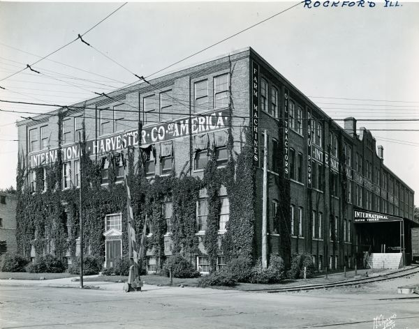 Exterior view of International Harvester's Rockford branch building. Railroad tracks pass a loading dock along the right side of the ivy-covered building and signs read: "Farm Machines. Oil Tractors. P&O Plows."