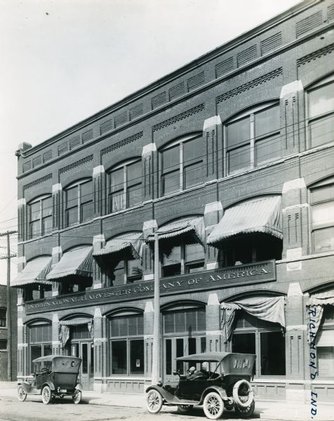 Exterior view of International Harvester's Richmond branch office. Two automobiles are park beneath awnings on the buildings facade, and a man is sitting waiting in the driver's seat of the automobile on the right.