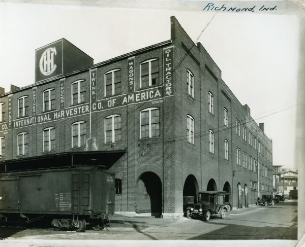 Exterior view of International Harvester's Richmond branch building. A train car rests on tracks that run adjacent to the building. Several automobiles are parked along and inside arched openings on the right side of the building.