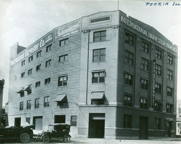 Exterior view of International Harvester's Peoria branch building. A sign along the top of the building advertises: "Home of International Motor Trucks." Two automobiles are parked along the street in front of the building.