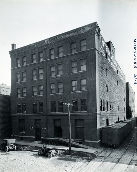 Elevated view of the front and side of an International Harvester branch office building in Milwaukee. Railroad cars rest on a railroad track that passes by the side of the building, and automobiles are parked along the curb in front. The numbers on either side of the front door read "217" and "225."