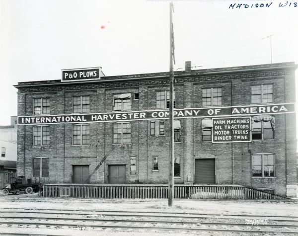 Exterior view of an International Harvester branch office building in Madison. Railroad tracks pass by the building and painted signs read, "P&O Plows. International Harvester Company of America. Farm Machines, Oil Tractors, Motor Trucks, Binder Twine."
