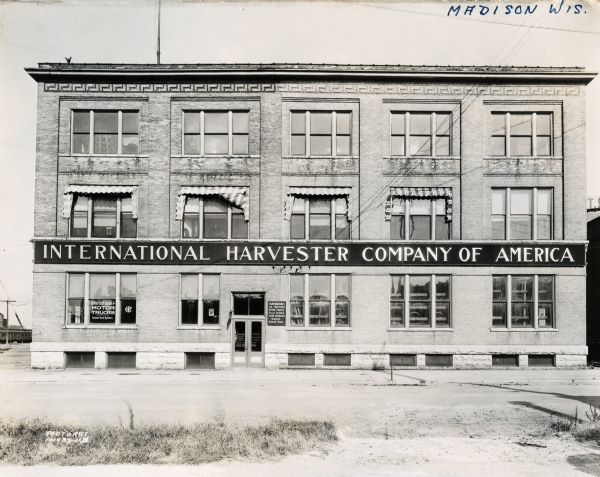 Exterior view of an International Harvester branch office building in Madison. The sign painted on the window reads, "International Motor Trucks. Exclusive Truck Builders" and the sign to the right of the entrance reads, "Farm Machines. Oil Tractors. Motor Trucks. P&O Plows. Cream Separators. Wagons. Binder Twine."