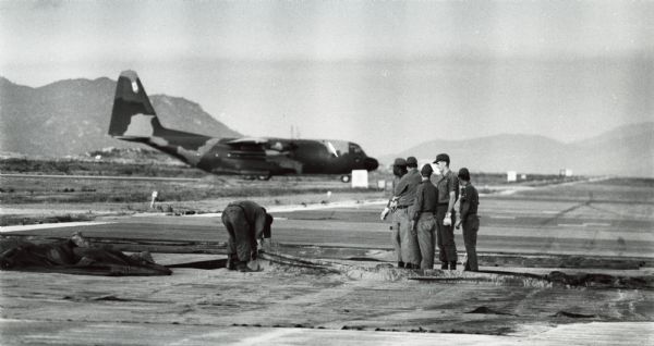 A group of men from the Air Force's Red Horse heavy repair squadrons stand outdoors while making repairs on an aluminum runway in Vietnam. An airplane is behind them, and in the background are mountains.