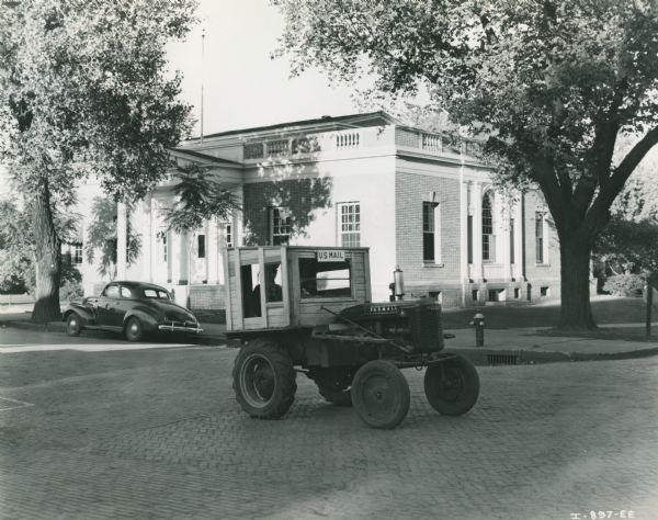 Farmall A tractor fitted with a homemade, wooden cab for the purposes of delivering mail, parked in front of a post office. A car is also parked in front of the building. The original caption reads: "T.D. Ashby, Chariton, Ia."