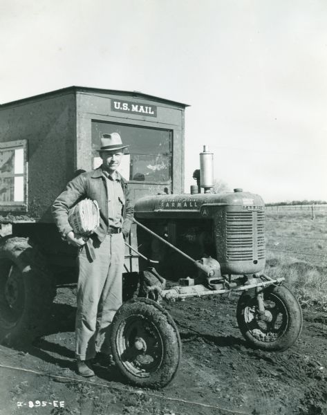 Levi Fisher stands holding a bundle of mail in front of a Farmall A tractor that has been fitted with a homemade cab to deliver the U.S. Mail. The cab has a decal that reads: "U.S. Mail." The original caption reads, "Meet Levi Fisher, introducing Farmall-A in the rural route mail service at Macksburg, Iowa."
