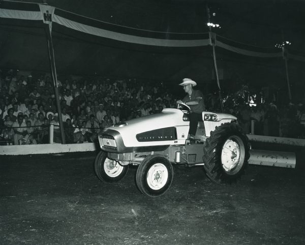 A man wearing a cowboy hat is operating an International Harvester model HT 340 experimental research tractor in a dirt arena under a tent in front of spectators.