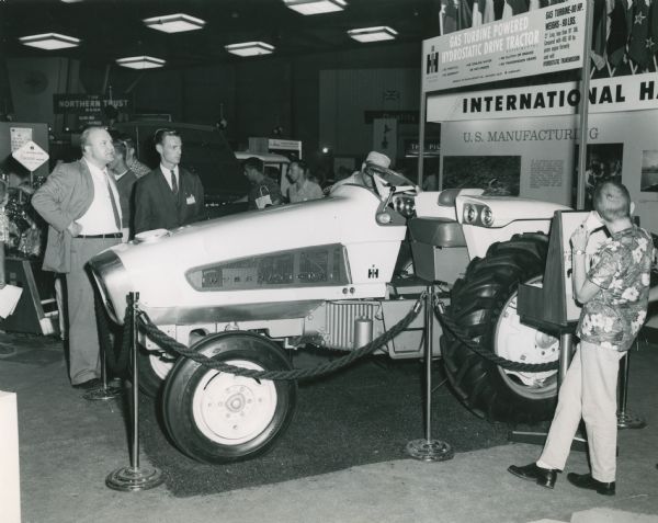 Men gather to look at an International Harvester Model HT-340 tractor on display behind a rope. The sign above the tractor reads, "Gas Turbine Powered Hydrostatic Drive Tractor. Experimental. Gas Turbine - 80 H.P. Weighs - 90 LBS. 21" Long, less than 19" DIA. Compared with 450, 40 hp. piston engine formerly used with Hydrostatic Transmission. No Throttle. No Gearshift. No Cooling Water or Anti-Freeze. No Clutch or Brakes. No Transmission Gears."