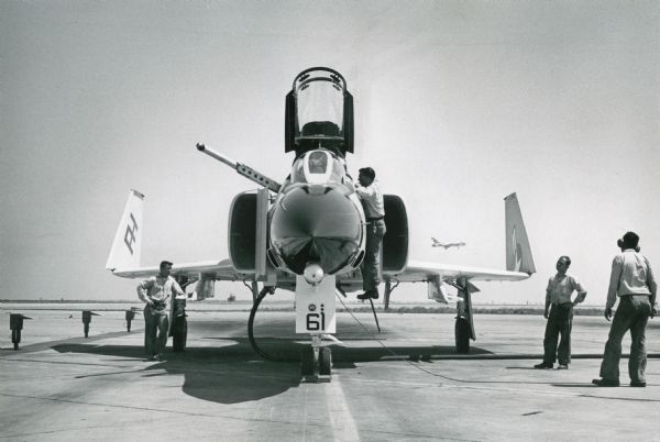 Front view of a Phantom II Naval plane on a tarmac at Miramar Naval Air Station. Men stand on and beside the airplane. Another plane is taking off in the background.