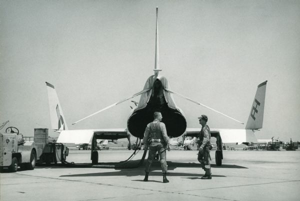 Rear view of a Phantom II Naval plane on a tarmac at Miramar Naval Air Station. Two uniformed men stand beside the airplane, and other equipment is in the background.