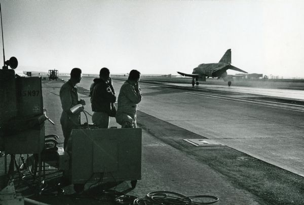 Three men stand alongside a tarmac as a Phantom II Naval plane takes off at Miramar Naval Air Station.