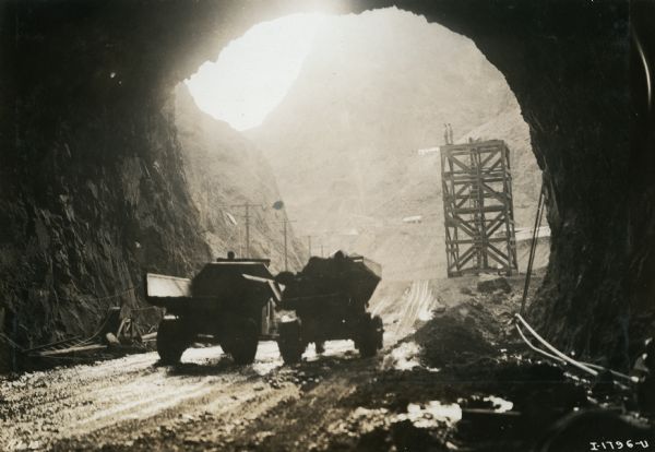 Two International trucks exit a tunnel on a dirt road leading through a canyon during the construction of the Hoover Dam. Two men stand on the top of a square tower near the tunnel.