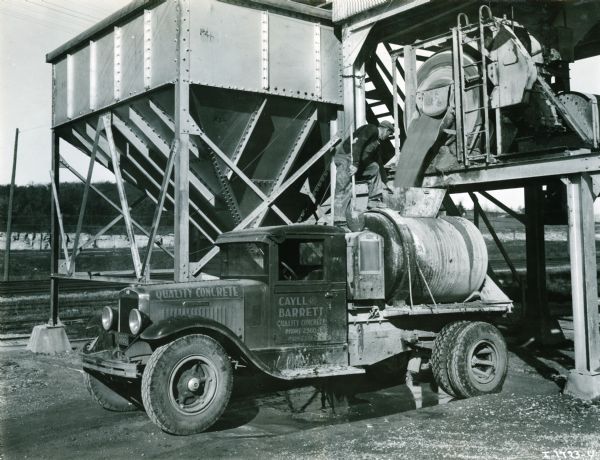 A man is standing on top of an International A-5 truck which is parked beneath a concrete spout to fill the tank on the truck bed.