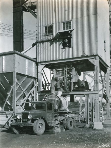 A man is standing on top of an International A-5 truck, filling a tank with concrete. A man wearing a hat is standing above him looking out of a window in a building.