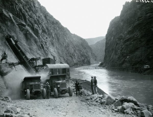 Men stand along a riverbank as International trucks are used to haul loads from the cliffs during the construction of the Hoover Dam. An electric shovel is working on the side of the road in the background.