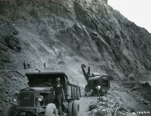 Men use International trucks to loads from the cliffs during the construction of the Hoover Dam. One of the men is smoking a cigarette. Two shovels are working in the background.