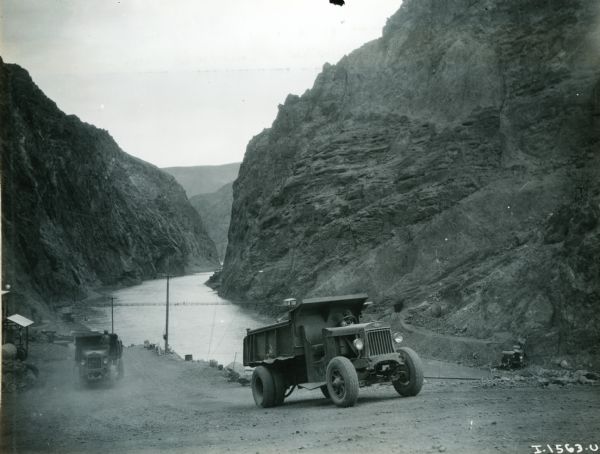 Two International trucks drive up a dirt road during the construction of the Hoover Dam.