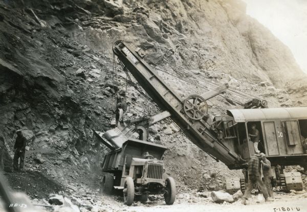 Men use dynamite, electric shovels, and an International truck to chip canyon walls down during the construction of the Hoover Dam.