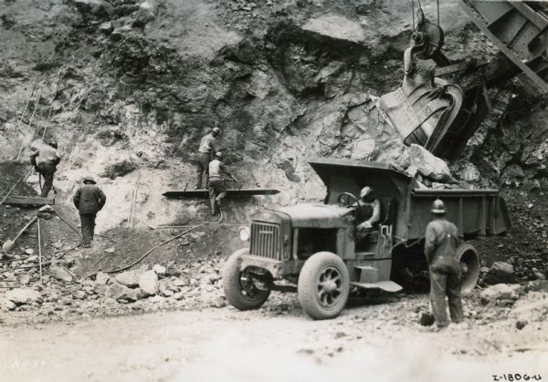 Men are standing on scaffolding built into the walls of a canyon during the construction of the Hoover Dam. An electric shovel is moving pieces of dynamite-blasted boulders into the bed of an International truck.