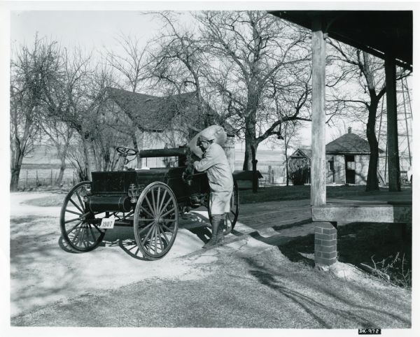 A man loads a burlap bag into an International Auto Wagon parked beside a farmhouse. Other farm buildings are in the background.