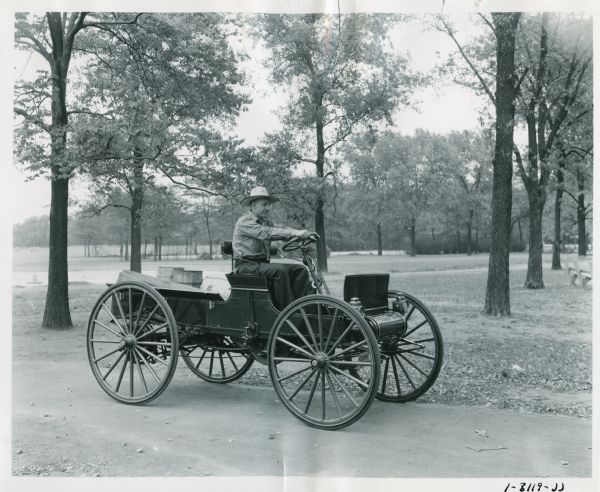 A man drives an International Auto Wagon along a dirt road through what appears to be a park.