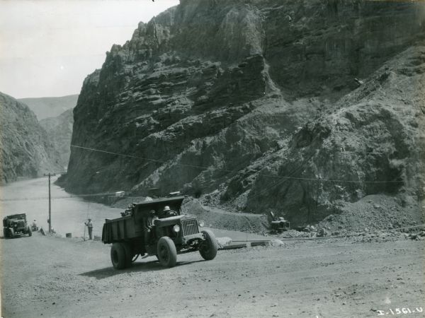 Men drive an International truck up a dirt road along the Colorado River during the construction of the Hoover Dam in Black Canyon. There is a steam, or power shovel on a road on the opposite bank of the river.