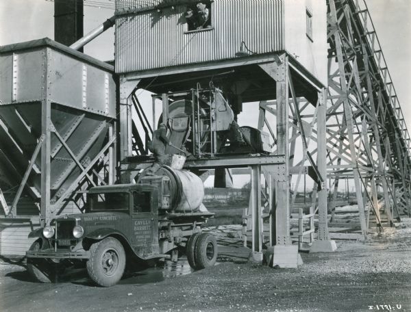 A man loading cement into the back of an International Model A-5 truck. The automobile is marked on the driver's side door with the words: "Cayll & Barrett Quality Concrete." Two men wearing hats are standing above him in the building over the spout looking out of a window.