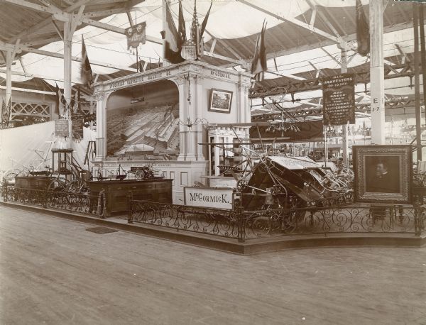 View of the McCormick display at the World's Columbian Exposition in Chicago (World's Fair of 1893). The exhibit features several full size and scale model agricultural machines, including a grain binder and a reaper. A large framed portrait of Cyrus Hall McCormick is on the right, and in the back of the exhibit, the McCormick Reaper Works factory in miniature.