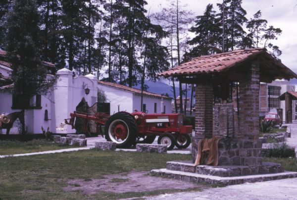 Buildings near International Harvester's Saltillo Works in Mexico. A donkey stands at left, and a tractor is parked in front of a building near a large, stone well with a tiled roof. The factory building is in the background.