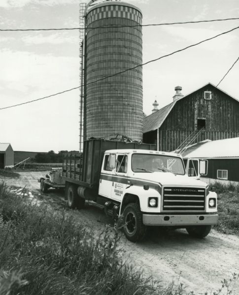An International S-Series truck with a trailer on a dirt road in front of a newly-constructed silo and several barns and farm buildings.
