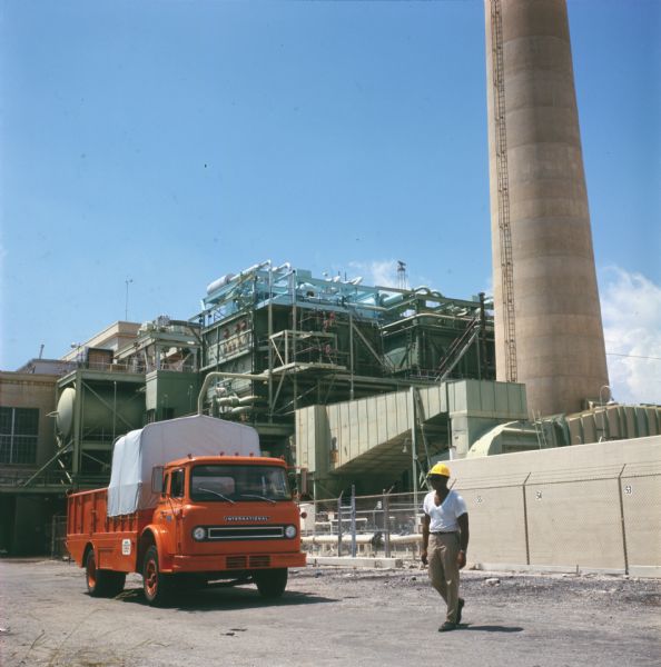 View of a man with a hard hat walking near an International truck parked in front of what appears to be a power plant. The truck is labeled with an "FPL" logo, which may stand for Florida Power and Light.