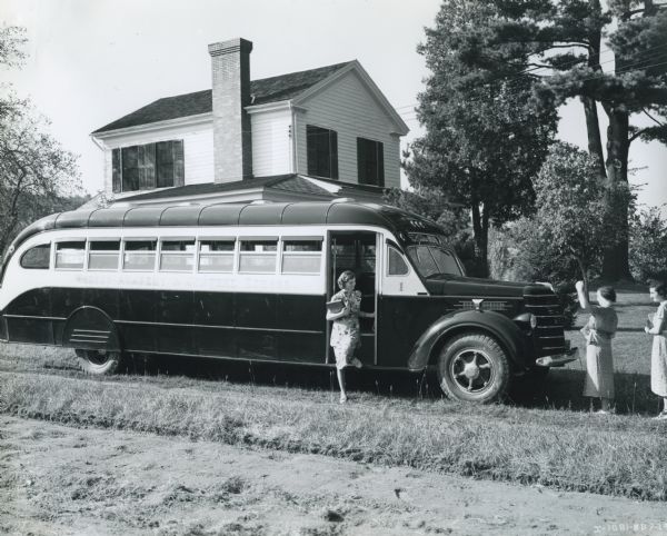 Side view of a woman disembarking from a 49-passenger International DS-40 bus. Two women stand near the front of the bus, and there is a house in the background. The bus was equipped with a 235-inch wheel base and Penn Fan Deluxe body.