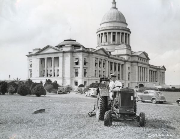 A man wearing a hat and tie is driving an International I-12 industrial tractor with mower attachment to groom the grounds in front of the Arkansas state capital. Original caption reads: "Capital grounds, Little Rock, Ark. I-12 tractor and mower being demonstrated."