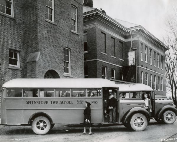 Greenfork Township School students aboard two International D-35 buses parked in front of two large, brick buildings. There appears to be an adult in front of one of the buses, possibly a teacher(?). One bus is labeled "Greenfork TWP. School, Randolph County, No. 6." These D-35 buses had 179-inch wheel bases and Union City Bodies.