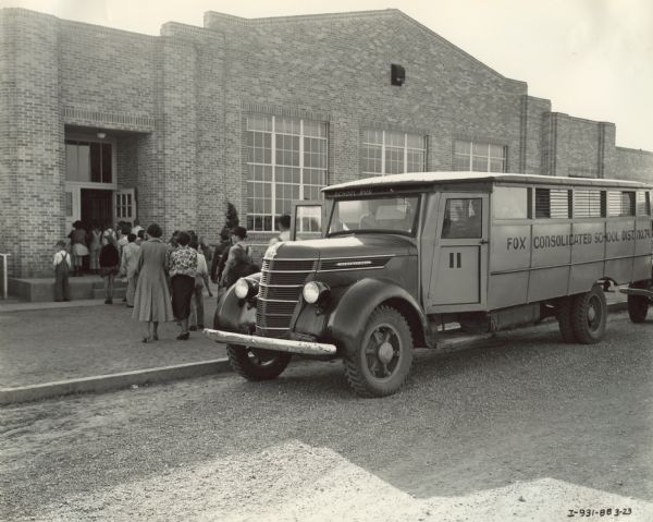 Students filing off of an International D-35 bus and into a school building, accompanied by a pair of adults, possibly teachers (?). The bus was equipped with a Picayune body, 179-inch wheel base. The side of the bus reads, "Fox Consolidated School Dist. No. 74."
