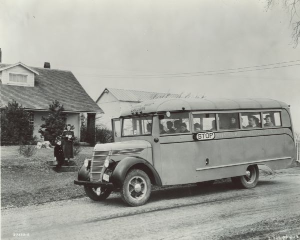 Two students approaching an International D-30 bus full of other children. The bus is parked on a residential street, slick with rain,  with houses in the background. The bus was equipped with a 155-inch wheel base and a 17 foot Wayne body.