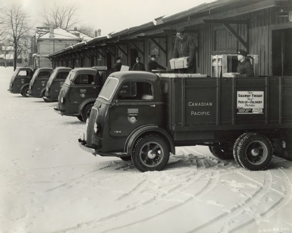 Five International D-300 trucks used by Canadian Pacific Express Company as delivery trucks for freight. There are men loading freight into the back of the trucks for delivery. Two of the trucks had 117-inch wheel bases. The other three trucks pictured had 99-inch wheel bases.